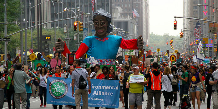 Protesters holding signs and banners walking down a New York City street. In the center of the crowd is a giant puppet. 