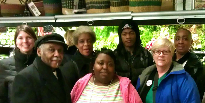 Seven people stand, smiling, facing the camera, in front of the vegetable section of a grocery store.